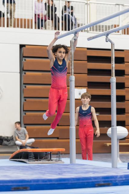 Young gymnast in pink outfit hanging from parallel bars while another child watches during training session in gymnasium