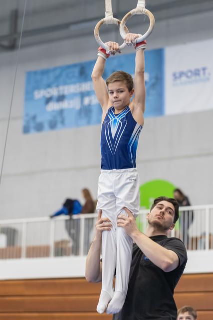Young gymnast in blue leotard hangs from rings with focused expression while coach spots from below in training facility