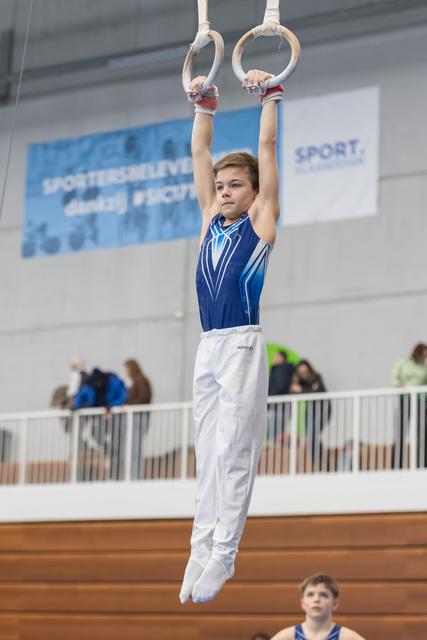Young gymnast in blue and white uniform hangs from still rings, focused expression, spectators visible in background