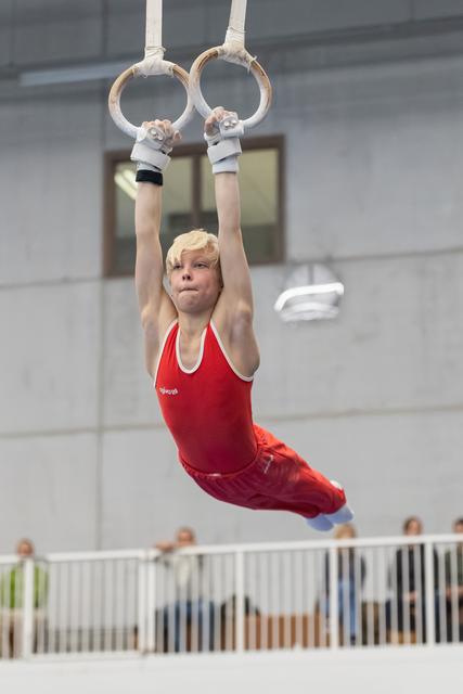 Young gymnast in red leotard hanging from rings with concentrated expression during routine at indoor gymnasium