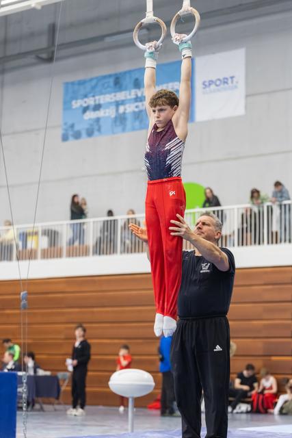 Young gymnast in red pants hangs from rings while coach spots below, demonstrating concentration during training routine