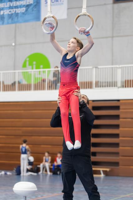 Young gymnast in red gradient leotard performs ring routine while coach spots from below in indoor training facility