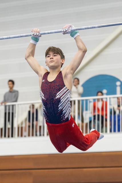 Young gymnast hanging from high bar with focused expression, wearing red and blue uniform with hand grips at indoor training facility