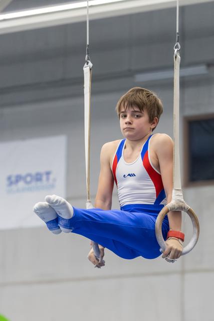 Boy gymnast holds L-sit position on rings, wearing blue and red uniform, demonstrating strength and focus in training gym