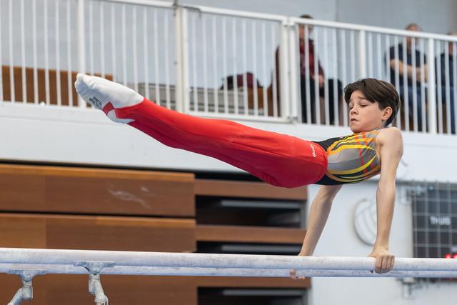 Young gymnast performs L-sit hold on horizontal bar with intense concentration, wearing colorful leotard and red pants