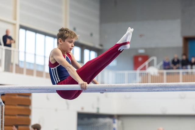 Young gymnast performs L-sit position on horizontal bar with extended legs, demonstrating strength and control in training facility