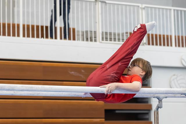 Young gymnast in red performs on horizontal bar, legs extended upward, in indoor gymnasium