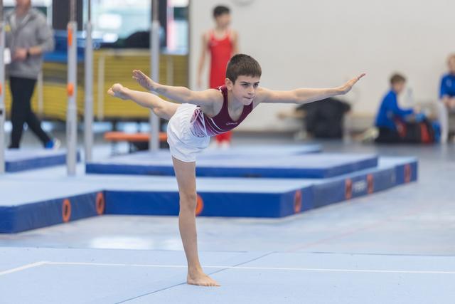Young gymnast performs a horizontal scale balance on floor, arms extended, demonstrating strength and control in training gym