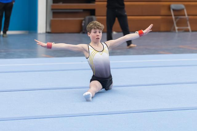 Young gymnast performs kneeling pose with arms extended during floor routine on blue mat in indoor training facility