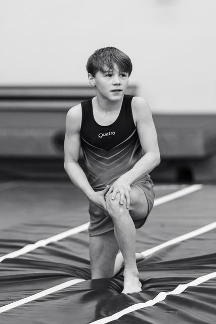 Young gymnast in black Quatro leotard kneels on one knee on striped floor mat, focused expression during routine