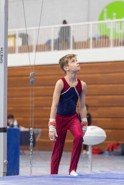 Young gymnast in red and navy leotard gazes upward after completing high bar routine, showing focused concentration
