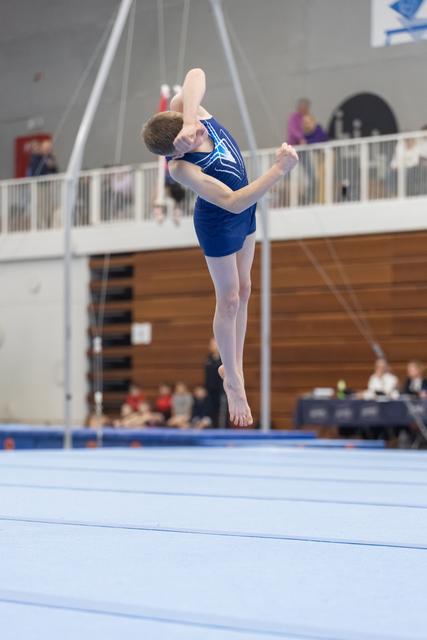 Young gymnast in blue leotard performs a high vertical jump with bent legs on floor exercise mat during routine
