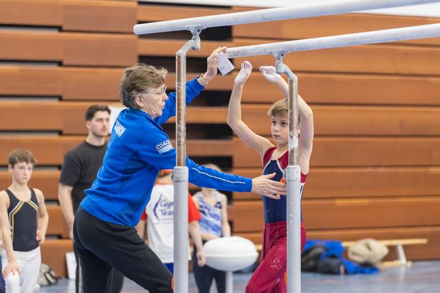 Young gymnast grips parallel bars while coach in blue adjusts equipment, wooden bleachers in background