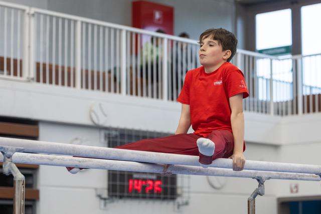Young gymnast in red uniform performs an L-sit position on parallel bars, displaying intense focus and determination