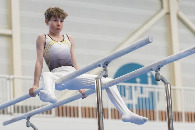 Young gymnast performs on parallel bars with focused expression, legs extended horizontally in white and blue uniform