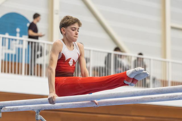 Young gymnast in red and white uniform performing L-sit position on parallel bars with concentrated expression