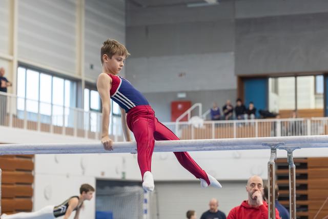 Young gymnast performs support hold on parallel bars with intense focus while spectators watch from the background