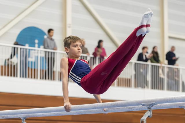Young gymnast performs an L-sit hold on parallel bars, demonstrating strength and balance while spectators watch from balcony
