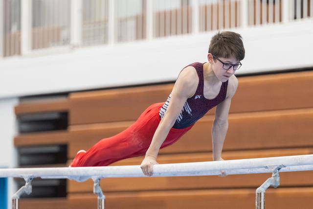Young gymnast performs on parallel bars, body extended horizontally, wearing glasses, purple leotard and red pants