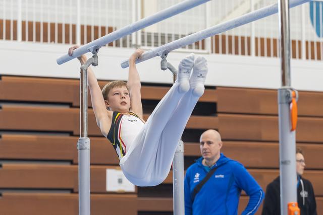 Young gymnast performs L-sit position on parallel bars while coach in blue jacket watches attentively from below