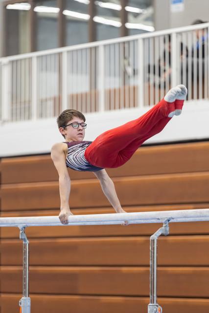 Young gymnast in glasses performing L-sit hold on parallel bars, demonstrating strength and focus in training facility