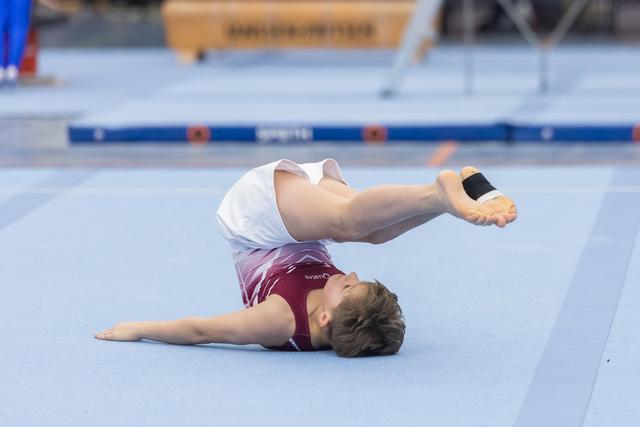 Young gymnast performs floor routine with back on mat and legs extended upward, demonstrating flexibility and control