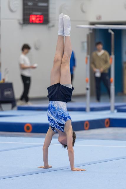 Young gymnast executes a handstand on blue mat, legs straight and pointed, while coaches observe in background