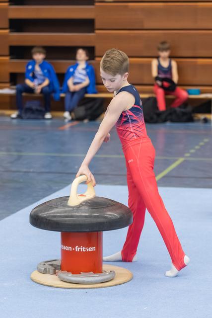 Young gymnast in red leotard practices mushroom circles while teammates watch from bleachers in indoor training facility