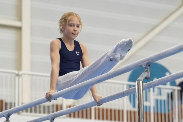 Young gymnast with blonde hair executes a move on parallel bars, legs extended upward in white pants and navy leotard