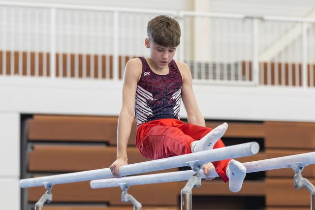 Young gymnast performs on parallel bars wearing red shorts and patterned leotard, focused expression in indoor training facility
