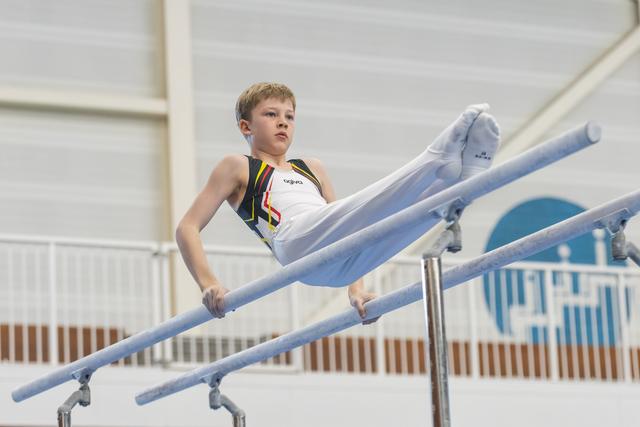 Young male gymnast executing a strength hold on parallel bars, legs extended horizontally, focused expression in training facility