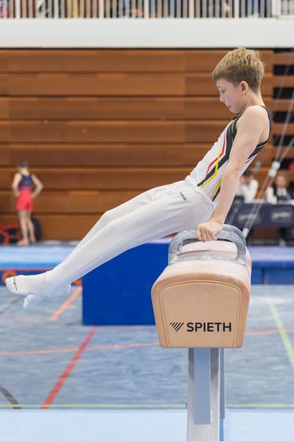 Young gymnast in white uniform executes a pommel horse routine with extended legs during indoor gymnastics meet
