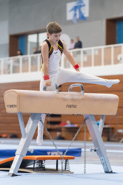 Young gymnast in white uniform executes a scissors move on pommel horse during routine, demonstrating form and concentration