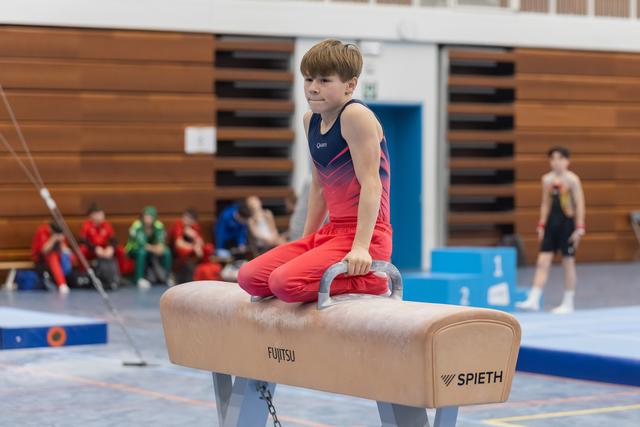 Young gymnast in red outfit executes pommel horse skill, focusing intently while teammates watch from background