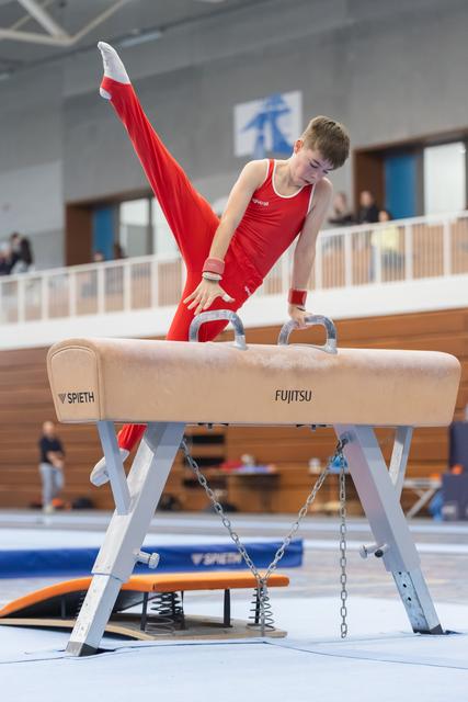 Young gymnast in red leotard executes a split-leg position on pommel horse, demonstrating strength and control in training facility