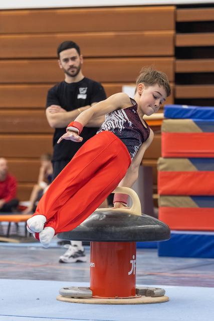 Young gymnast in red pants executes a scissors move on pommel horse while coach observes in gym setting