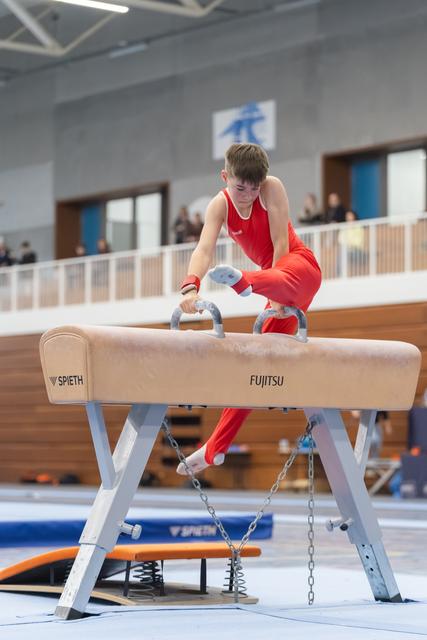 Young gymnast in red uniform concentrates intensely while performing on the pommel horse during an indoor gymnastics event