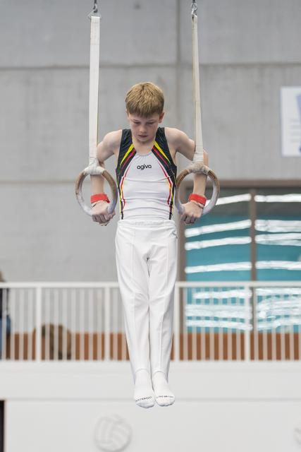 Young gymnast in white uniform performs rings routine with focused concentration at indoor training facility