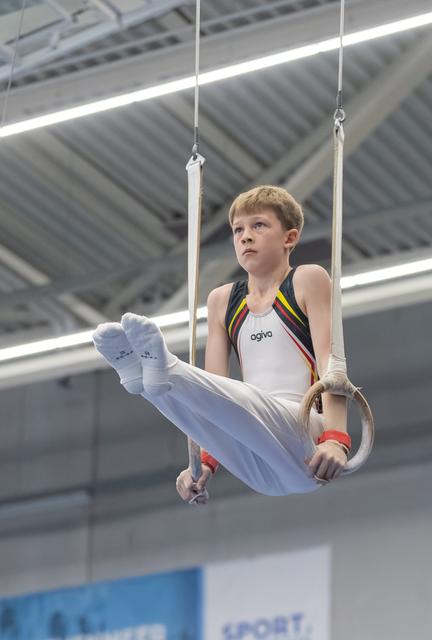 Young gymnast in rainbow-striped leotard executing an L-sit position on still rings with concentrated expression in indoor facility
