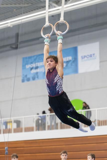 Young gymnast in patterned leotard performs on still rings with focused expression, body extended horizontally in gymnasium