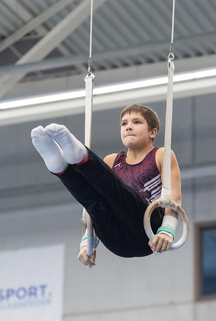 Young male gymnast concentrates while performing on still rings, legs extended upward in an indoor training facility