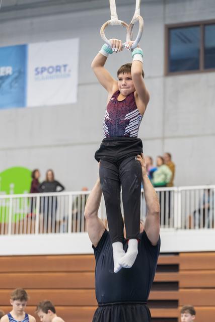 Young gymnast holds rings overhead while balanced on spotter's shoulders, displaying focus and concentration in indoor venue