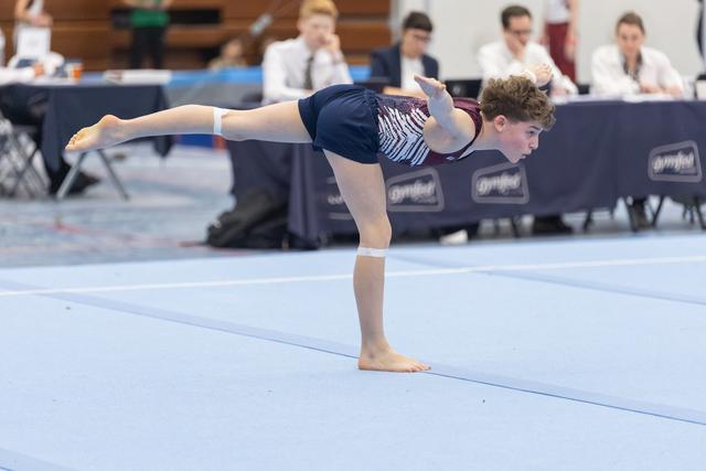 Young gymnast demonstrates a standing scale balance on floor mat, extending leg behind while judges observe from table