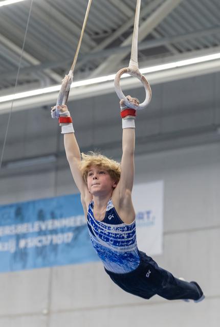 Young male gymnast with curly blonde hair holds a still position on rings, arms extended overhead in indoor training facility