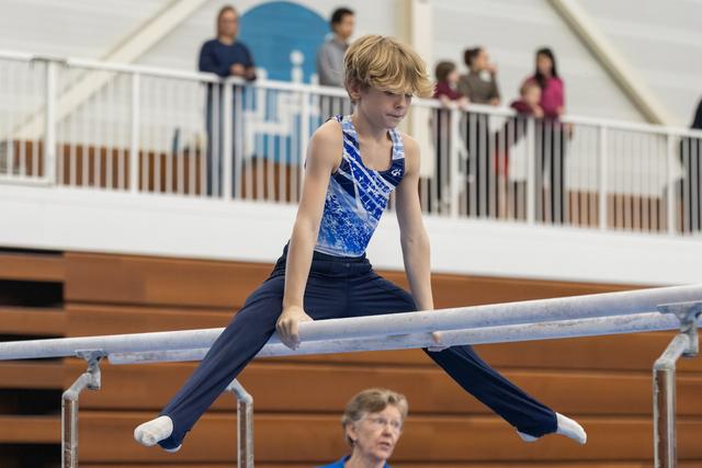 Young gymnast in blue leotard performs straddle position on parallel bars, focused expression, with coach below