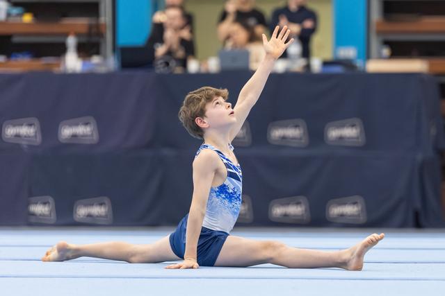 Young gymnast performs full splits on floor with dramatic arm extension, showcasing flexibility and artistic expression