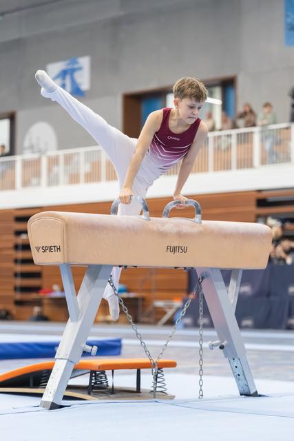 Young gymnast performs pommel horse routine with focused expression, legs extended upward in white-walled training facility