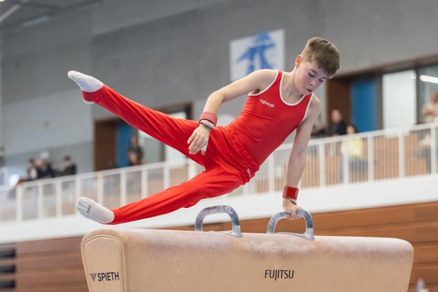 Young male gymnast in red uniform performs horizontal hold on pommel horse with legs extended, demonstrating strength and control