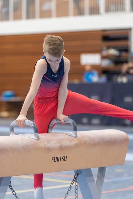 Young gymnast in red outfit performs on pommel horse with focused concentration during training session