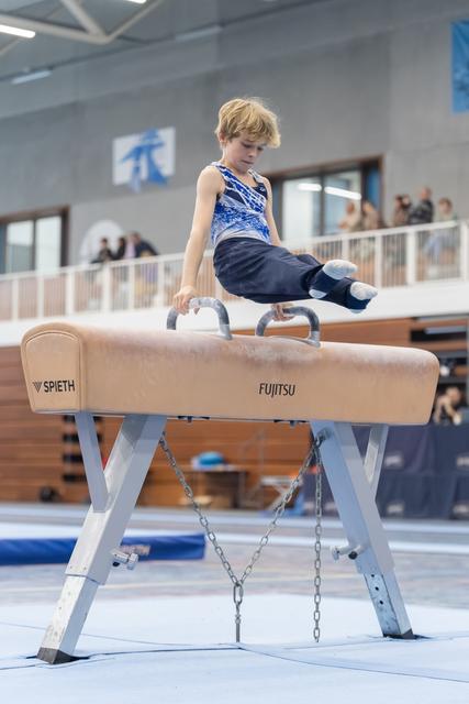 Young boy performs on pommel horse with focused concentration, wearing blue gymnastics attire in training facility